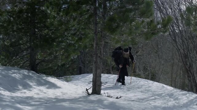 Orthodox monk trekking with a backpack and sticks on a snowy path in a forest on Mount Athos
