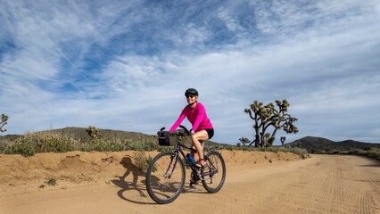 Fototapeta premium Mature woman rides her bike along a dirt path in Joshua Tree National Park surrounded by unique trees and open land under a cloudy sky
