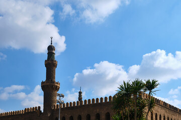 Obraz premium Low-angle view of historic mosque minaret rising above traditional Islamic architecture, framed by palm trees under bright blue sky with scattered clouds