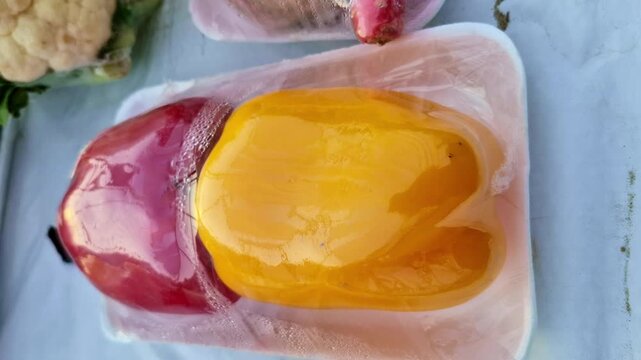 Red and yellow bell peppers (Capsicum annuum) placed on a tray and wrapped in plastic with condensation, displayed as fresh vegetables at a produce stall.
