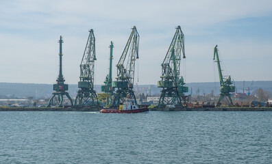 Obraz premium Industrial sea port landscape with green cargo cranes and a small red tugboat navigating the blue water under a clear sky. Commercial harbor infrastructure and shipping