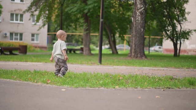 Toddler walking along park path, chasing colorful toy rolling ahead, tentative first steps on pavement, balancing near grass edge, neighborhood buildings blurred, active lifestyle scene, playful