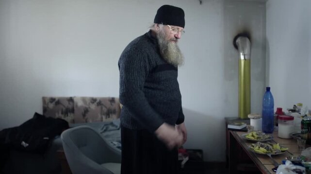 Elderly orthodox monk with a long beard arranging furniture inside his monastic cell in Greece