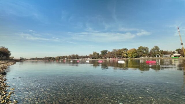 Timelapse view of calm water and colorful autumn trees around Sava Lake at Ada Ciganlija in Belgrade. Concept of seasonal landscape, urban nature, recreation and peaceful waterfront atmosphere