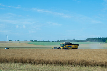 Fototapeta premium Yellow combine harvester working in a large golden wheat field with motion blur in the foreground and a wind turbine in the background