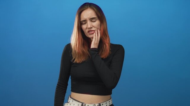 Woman touching cheek with hand showing toothache in studio, redhead wearing black top and patterned pants; discomfort worry.