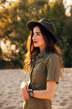 Young woman stands on sandy ground wearing a hat and a green shirt near trees during daytime