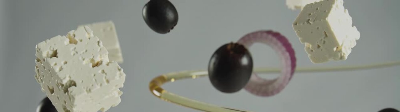 Feta cheese cubes, black olives, and red onion rings dispersing in the air, creating a dynamic food composition.