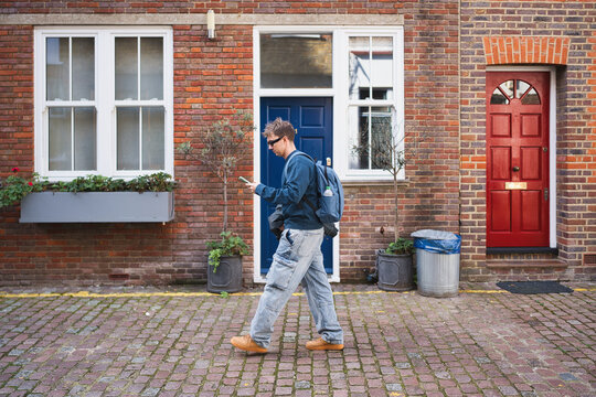Man in casual clothing and backpack walking on a historic london street, focusing on his mobile phone with traditional brick buildings in the background