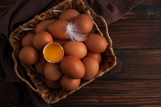 Rustic woven basket filled with fresh brown eggs, placed on wooden background with yalk into shall and delicate white feather resting on top