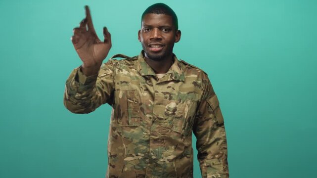 Soldier man in camouflage uniform waving hand in studio with teal wall and smiling face; duty welcome reassurance solidarity.