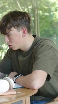 Vertical video: Leaning teen studying notebook, resting hand on temple at home window, in olive tee