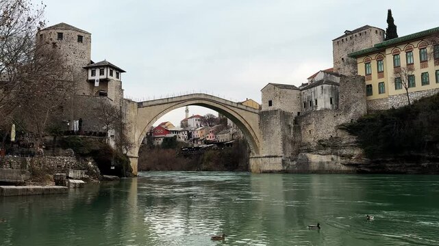 Daytime view of the historic Stari Most stone bridge over emerald river in Mostar with old town buildings and ducks on calm water. Iconic Balkan landmark showing heritage, culture