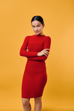 Young woman in red dress stands with crossed arms looking confidently at camera on yellow background. Female model with dark hair in neat style wears tight fitted outfit for fashion portrait.
