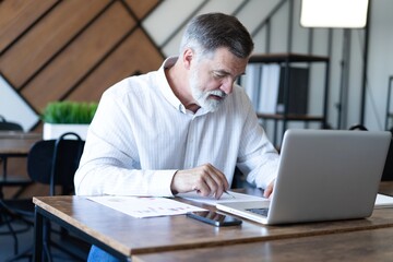 Fototapeta premium Smiling mature business man executive wearing shirt sitting at desk using laptop. Happy busy professional middle aged businessman investor working on computer looking away in office