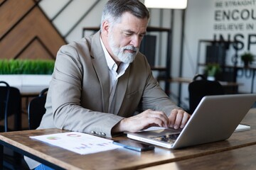 Fototapeta premium Smiling mature business man executive wearing shirt sitting at desk using laptop. Happy busy professional middle aged businessman investor working on computer looking away in office