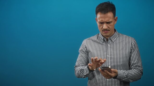 Man tapping smartphone with index finger while holding phone in hands against blue backdrop in studio; frustration concentration.