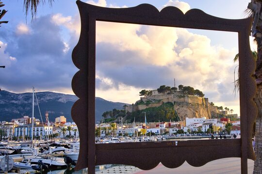 Framed view of the marina harbor and historic castle hill in D&eacute;nia, Spain at golden sunset. Sailboats and waterfront promenade create a cinematic Mediterranean coastal cityscape under dramatic clouds.