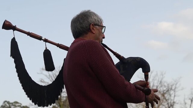 A profile shot of an elderly man with glasses playing the bagpipes against a bright, cloudy sky, showcasing the full instrument and its fringes.