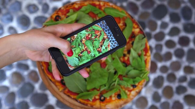 Close up top down perspective of a woman's hand taking a picture of a fresh vegetarian pizza with her mobile phone for social media sharing, capturing the perfect shot for her food blog