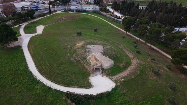 Close-up aerial shot of Dolmen de Menga, a massive megalithic burial site in Antequera, Spain. The drone flies closely over the grassy mound and stone entrance, revealing its size and alignment. 