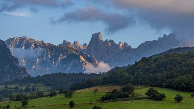 Majestuosa vista del Naranjo de Bulnes en los Picos de Europa, con sus paredes calizas iluminadas por el sol poniente sobre un valle verde y bosques envueltos en jirones de niebla.