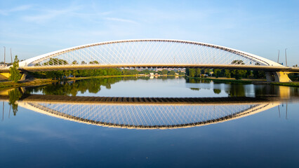Troja Bridge stands over the Vltava River in Prague during sunrise. The calm waters reflect the structure, creating a clear view of the bridge and surroundings. © pyty