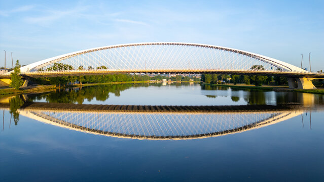 Troja Bridge stands over the Vltava River in Prague during sunrise. The calm waters reflect the structure, creating a clear view of the bridge and surroundings.