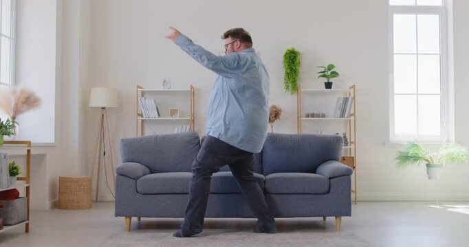 Expressive fat overweight man in glasses dancing and singing with upbeat energy near a sofa in living room, enjoying music and the moment at home, carefree. Obese male person having fun indoors.