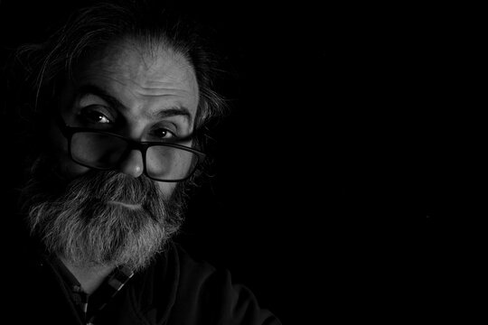 Dramatic Black and White Portrait of Thoughtful Bearded Man in Low Key Lighting Moody low key black and white portrait of a mature bearded man resting his head on his hand, looking at the camera with 