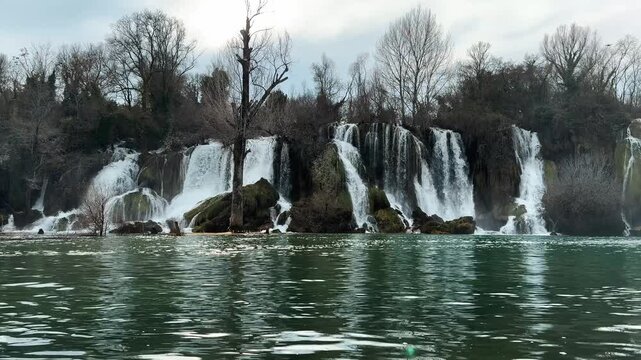Wide view of Kravica Waterfall with multiple cascades flowing into calm green river. Powerful natural landscape with forest, rocks, and moving water, ideal for travel, nature, and cinematic footage
