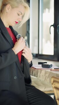 Young businesswoman preparing for live stream, attaching clip-on lavalier microphone to formal jacket and check smartphone before start recording session or video call.