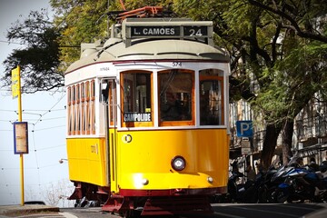 Naklejka premium Iconic yellow tram climbing through colorful houses in Lisbon, Portugal on a bright sunny day. Panoramic view of the city center with terracotta tiled roofs, blue sky and vibrant facades – classic Lis