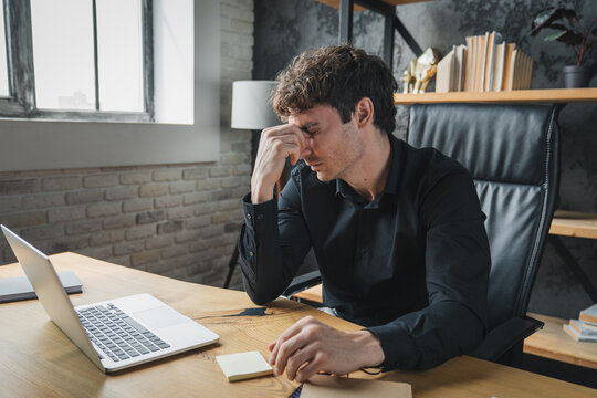 Tired businessman taking off glasses, massaging nose bridge leaning back in chair at workplace, overworked man suffering from computer vision syndrome after long laptop use, eye strain problem