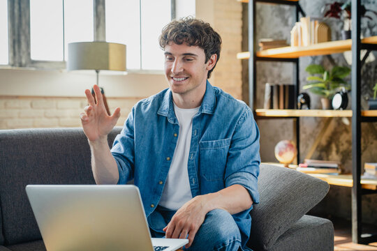 Happy young man makes videocall seated on sofa at home, greets friend or family start video conference using application on laptop. Virtual meeting event, tech, remote talk concept