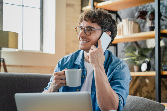 Remote work, laptop, phone and man on sofa with drink, conversation at home. Male person, small business and technology in living room for contact, ecommerce and interior design on internet