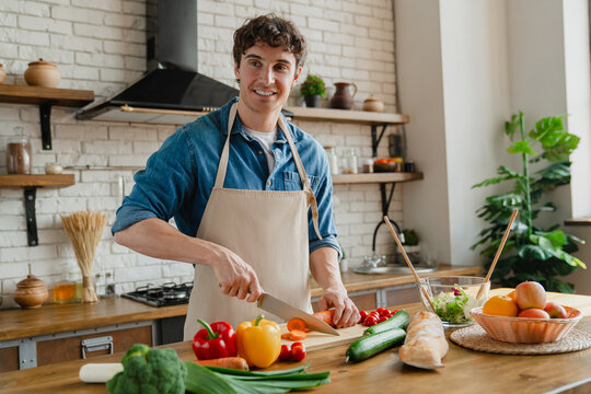 Happy young Caucasian chef man enjoying cooking hobby in contemporary home kitchen, chopping fresh vegetables on boars, preparing salad, smiling, laughing with appliance in background