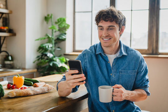 Handsome millennial single man cooking in modern kitchen holds smart phone searching recipes on internet, looking at device screen smile make food products delivery order, enjoy easy online services