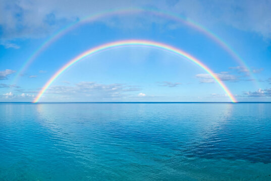 A vibrant double rainbow arches across a clear blue sky over a calm turquoise ocean