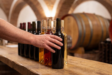 Naklejka premium Hand reaching for dark wine bottle on wooden table in cellar with barrel background