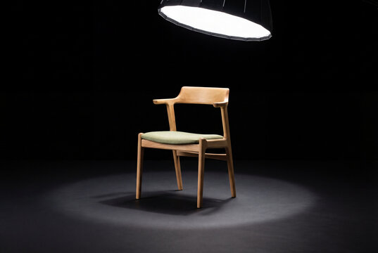 A single wooden chair with a green seat under a spotlight on a dark stage for an interview