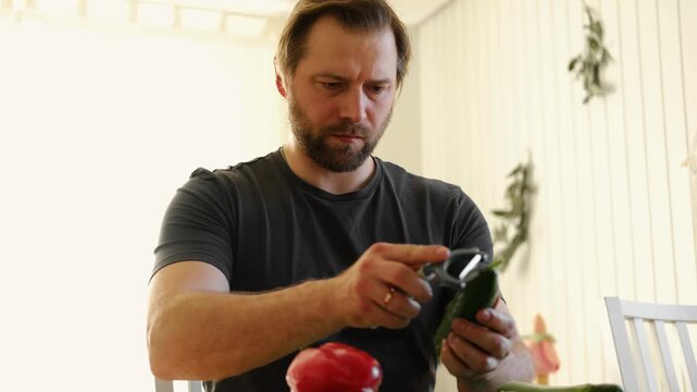 Man preparing fresh vegetables in modern kitchen, organic ingredients. Healthy meal. Cooking Food At Home. Male slicing red pepper and green cucumber on a wooden cutting board. Nutritionist, culinary