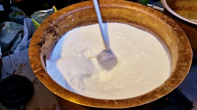 Large brass pot of tea and milk being stirred while simmering to prepare a traditional Indian dairy dessert at a food stall.