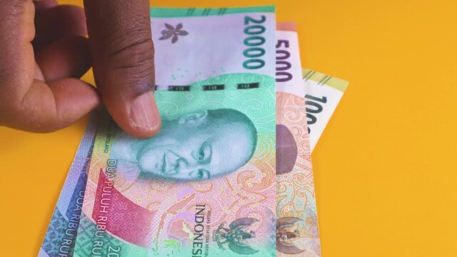 A hand places a series of Indonesian Rupiah banknotes on a yellow background, featuring denominations from 1,000 to 100,000, including the special 75,000 note, in a neat, overlapping arrangement.
