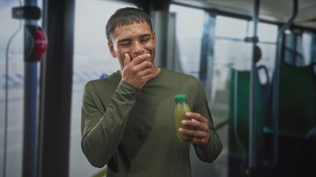 Young hispanic man holding a green capped juice bottle, hand over mouth smiling and laughing while standing on an airport shuttle bus near terminal windows; playful joy.