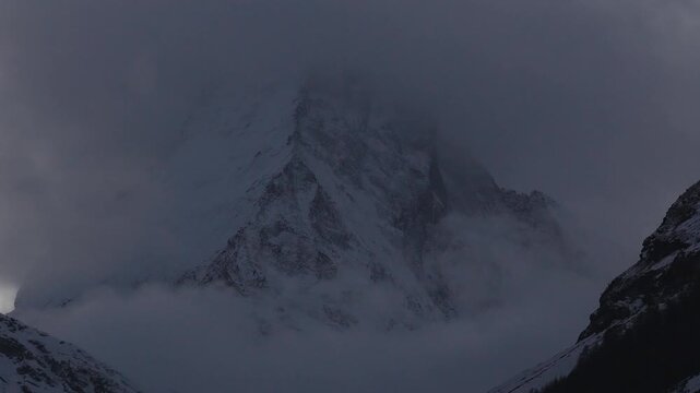 Aerial view of the Matterhorn above Zermatt, Switzerland, as clouds and fog drift over steep jagged faces, snowy ridgelines, and a dark treeline in cool low light.