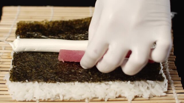 Professional sushi chef preparing a delicious maki roll with fresh raw tuna, cream cheese, and rice on a nori seaweed sheet placed over a traditional bamboo mat