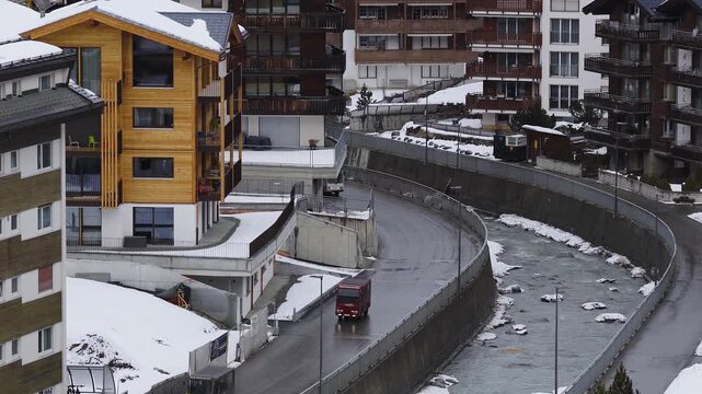 Aerial view of Zermatt, Switzerland, with timber chalets and curved flume as river flows, minibuses move on wet road, soft overcast light and tight composition