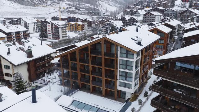 Aerial view over Zermatt, Switzerland shows snow, a timber and glass alpine building, chalets, terraced roofs, winding streets, light traffic, and a distant crane.