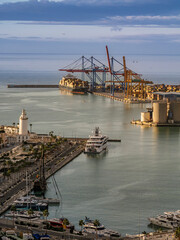 Fototapeta premium A panoramic view of a Mediterranean port at sunset featuring large container ship loaded by cranes, marina with luxury yachts, white lighthouse and waterfront industrial facilities, Malaga, Spain